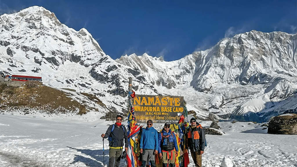 Trekkers standing in front of a “Namaste Annapurna Base Camp” sign, surrounded by snow-covered Himalayan mountains in Nepal.