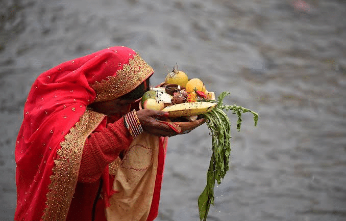 Devotee in red saree holding a bamboo tray of fruit offerings during Chhath Puja rituals.