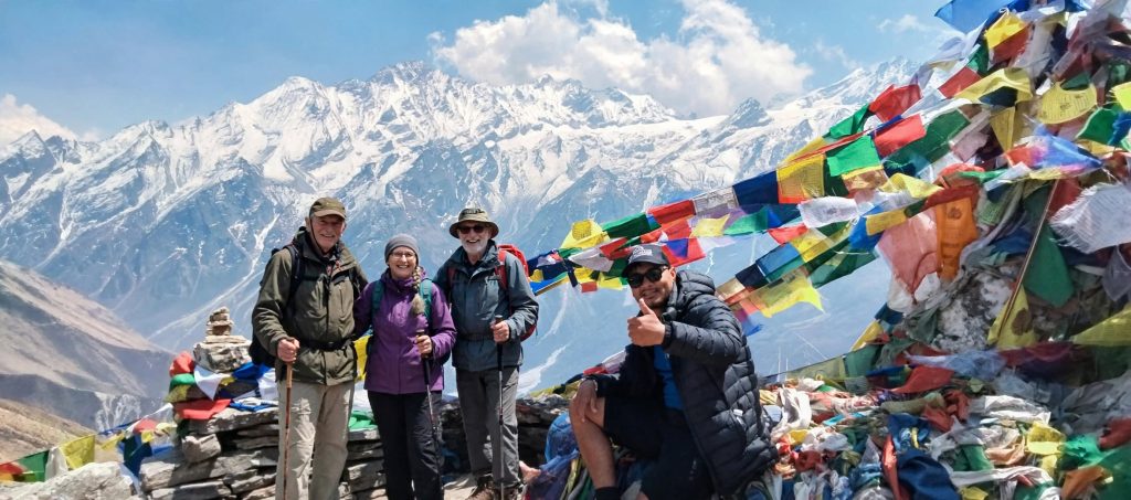 A trekking group resting on a ridge with prayer flags and layered Himalayan peaks behind them